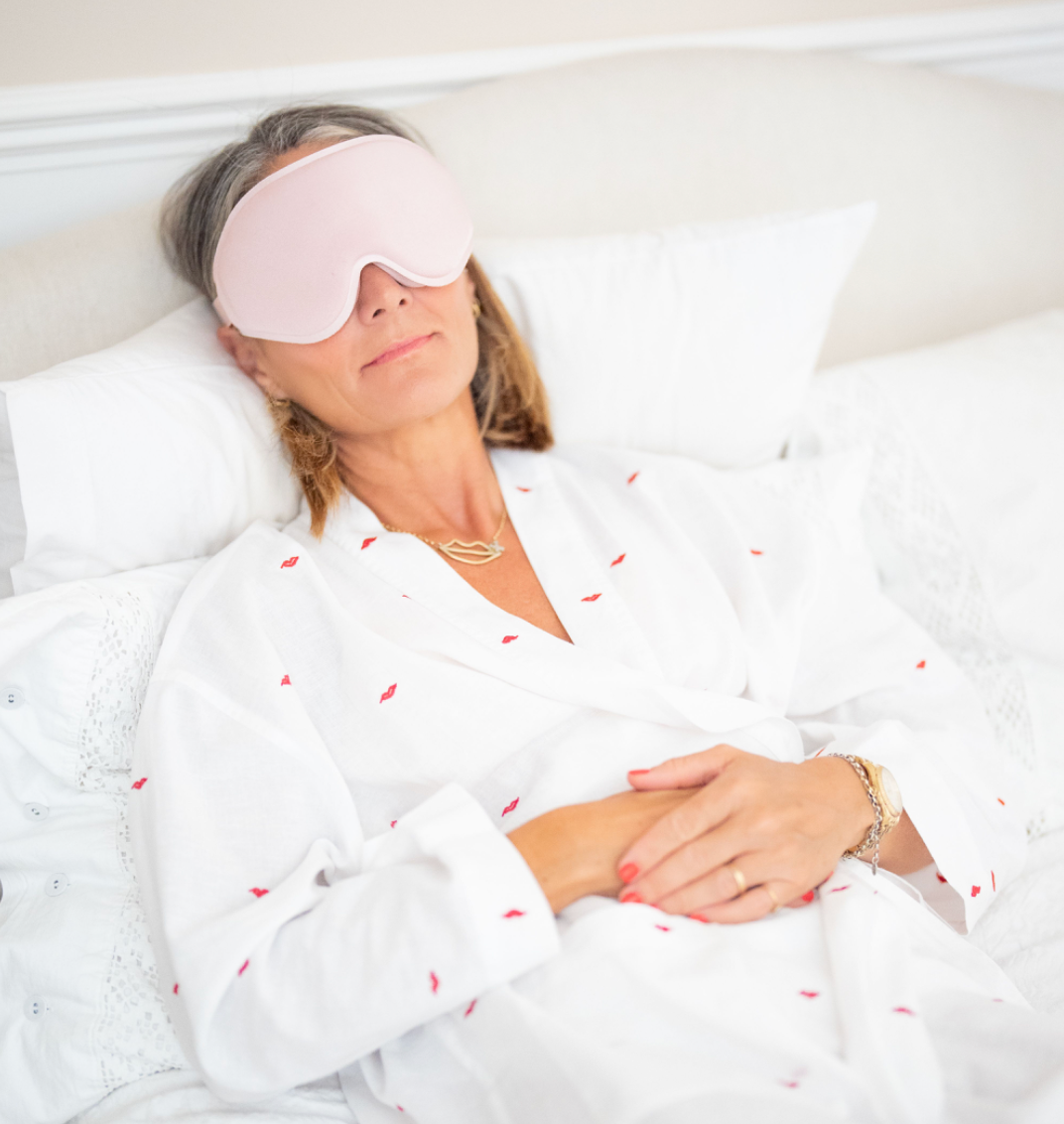 A woman lying in bed with white pillows and bedding, wearing a light pink sleep mask and a white robe with small red embroidered details.