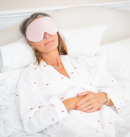 A woman lying in bed with white pillows and bedding, wearing a light pink sleep mask and a white robe with small red embroidered details.